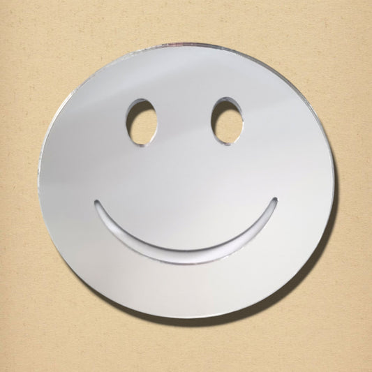 A round, silver smiley face mirror with oval eye cutouts and an upturned smile cut against a beige background.