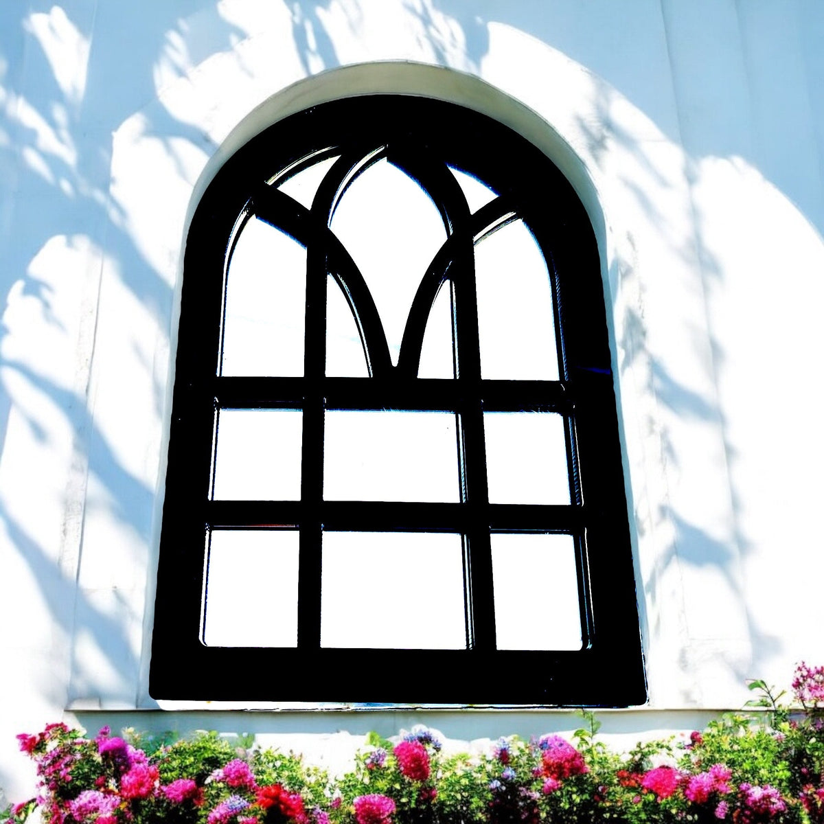 Black Victorian arch garden mirror mounted on a white wall above pink flowers.