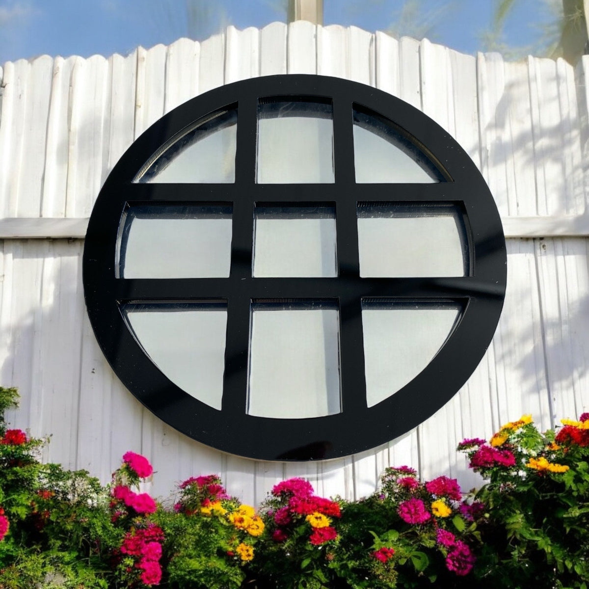 Round black framed garden mirror with a crossbar grid, mounted on a white wall above flowers.