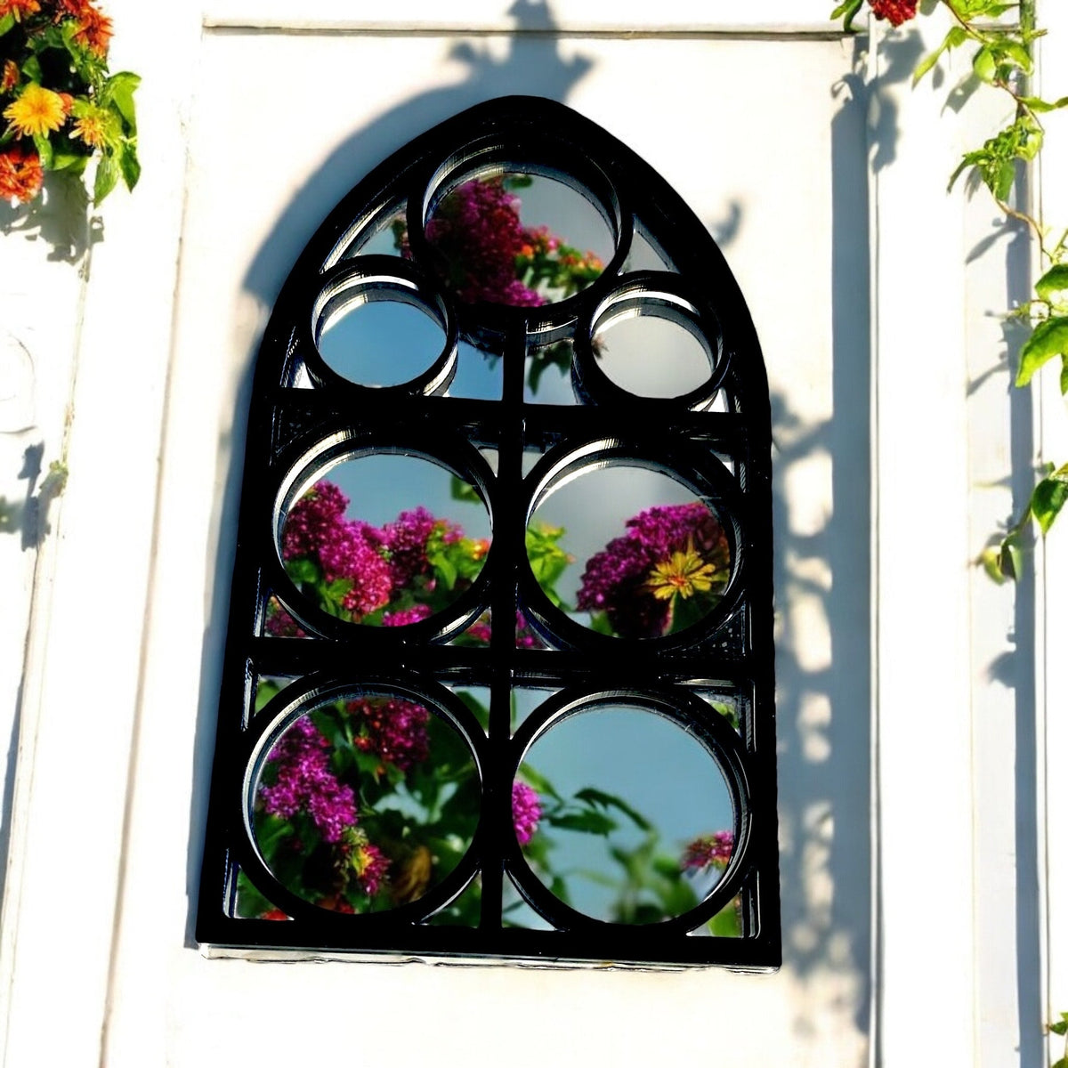 Black Gothic arch mirror with circular panes reflecting flowers, mounted on a white wall.