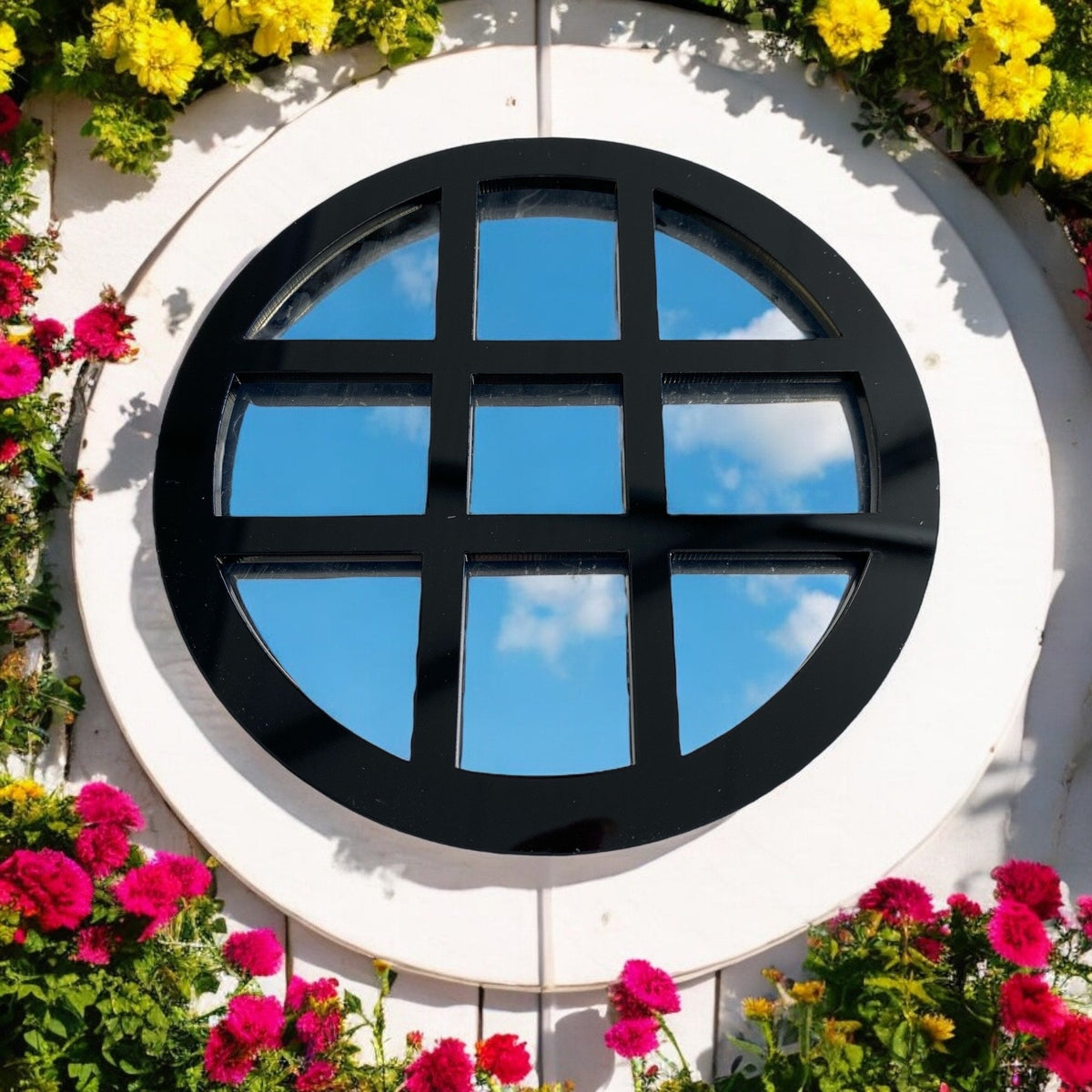 Round black framed garden mirror with cross bars, reflecting a blue sky with clouds, mounted on a white wall.