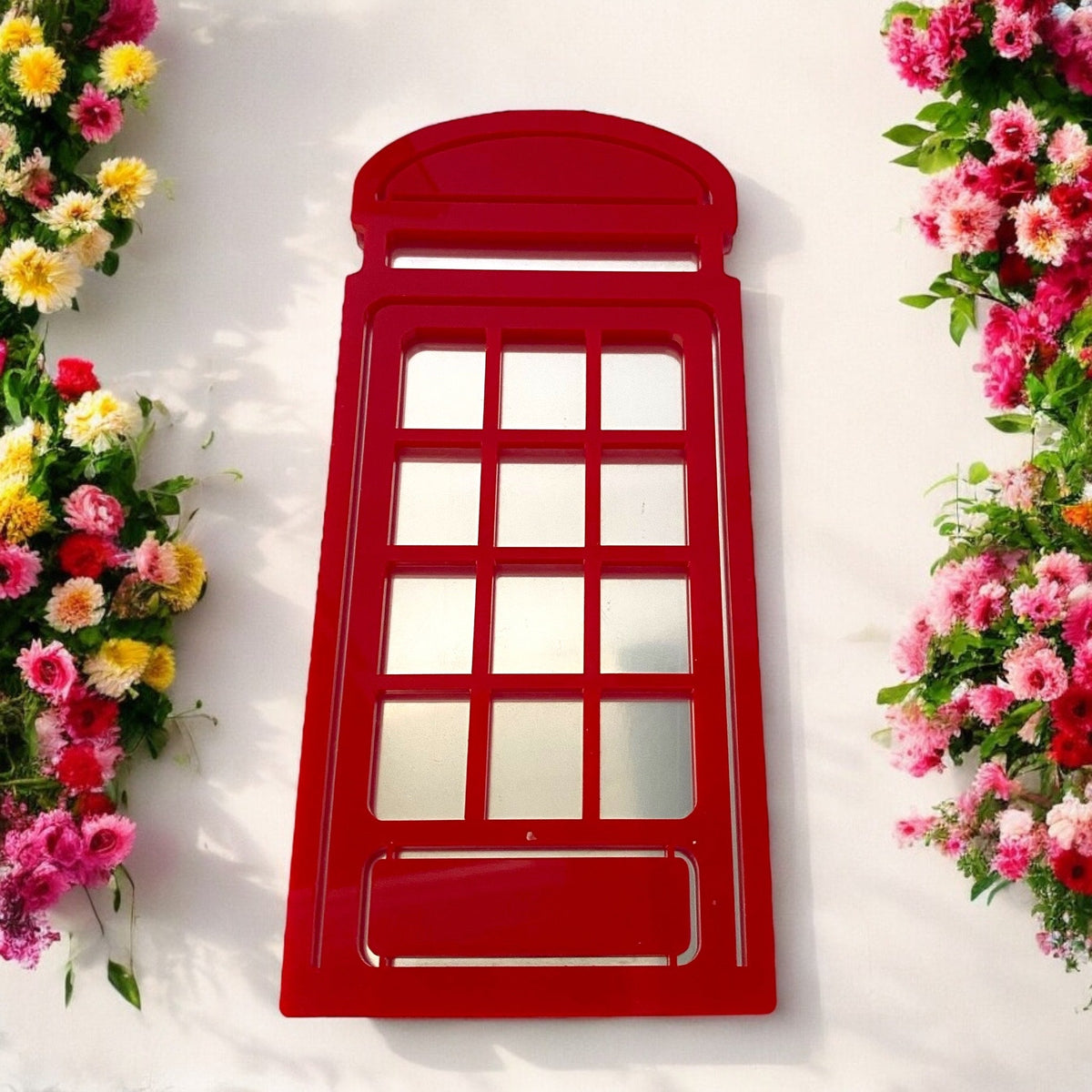 Red telephone box garden mirror mounted on a white wall between pink and yellow flowers.