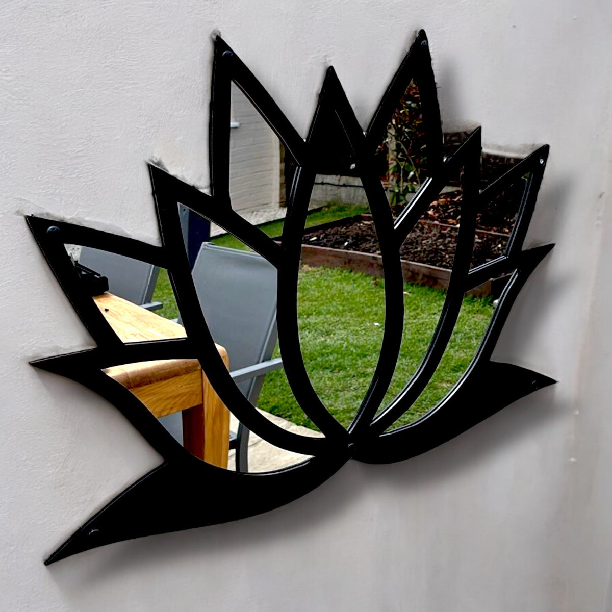 Black metal-framed lotus flower garden mirror mounted on a white wall, reflecting a patio and lawn.