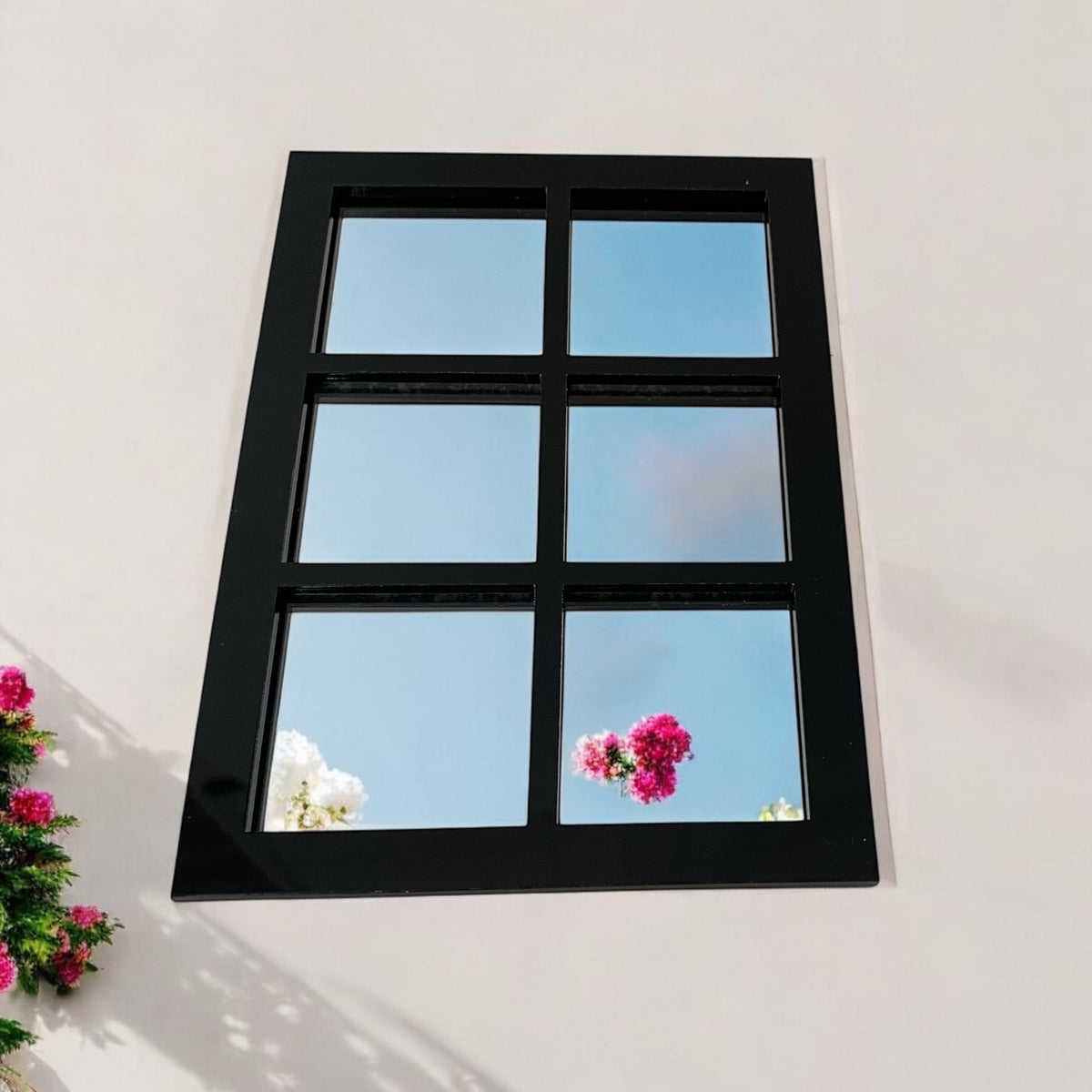 Black six-panel garden mirror mounted on an outdoor wall, reflecting the sky and pink flowers.