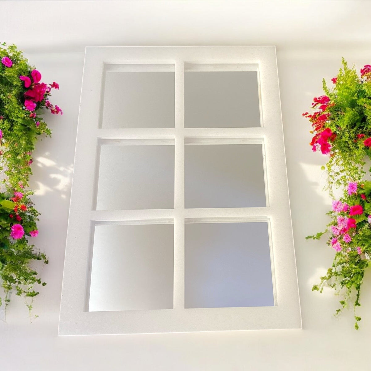 A rectangular white six-panel garden mirror mounted on a white wall, flanked by hanging planters with green foliage and pink flowers.