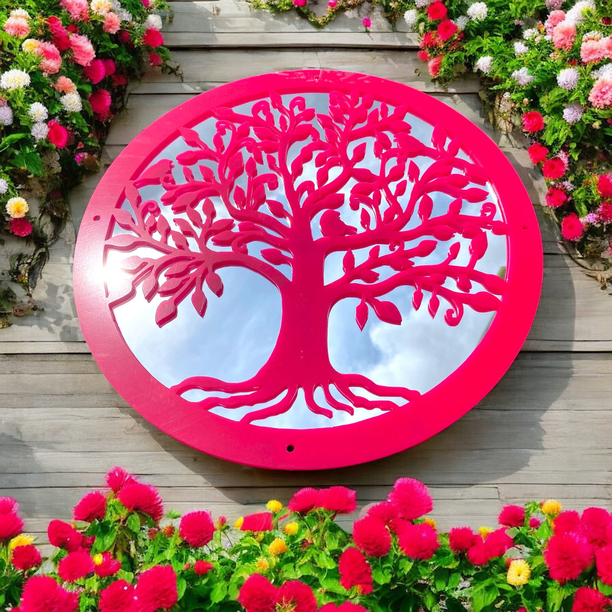 Round pink Tree of Life garden mirror with an ornate tree overlay, mounted on a wooden wall amid flowers.
