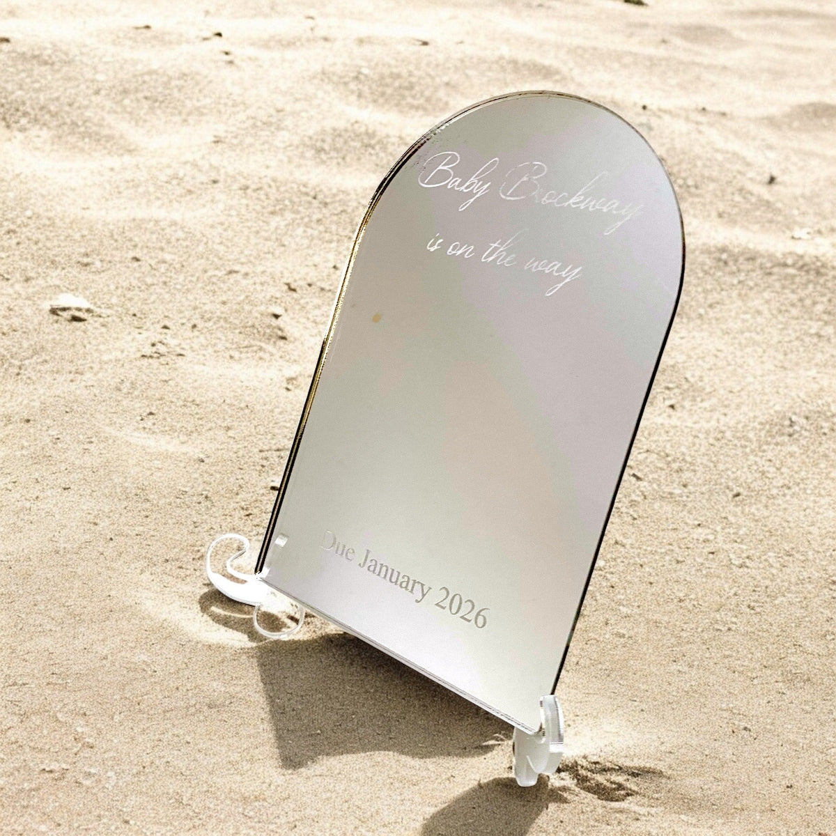 Custom arched mirror on a clear acrylic stand displaying engraved text "Baby Brockway is on the way Due January 2026" on sand.