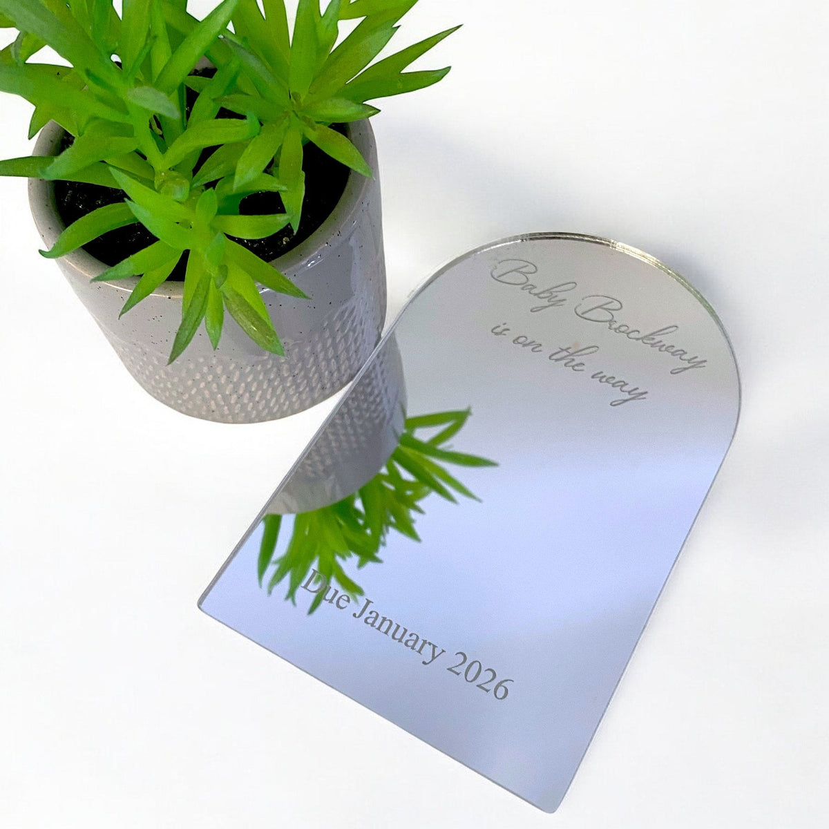 Custom engraved arch mirror with "Baby on the way" text, next to a small potted plant on a white surface.
