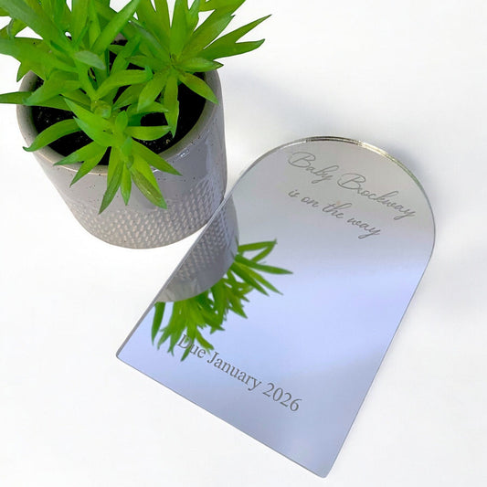Custom engraved arch mirror with "Baby on the way" text, next to a small potted plant on a white surface.