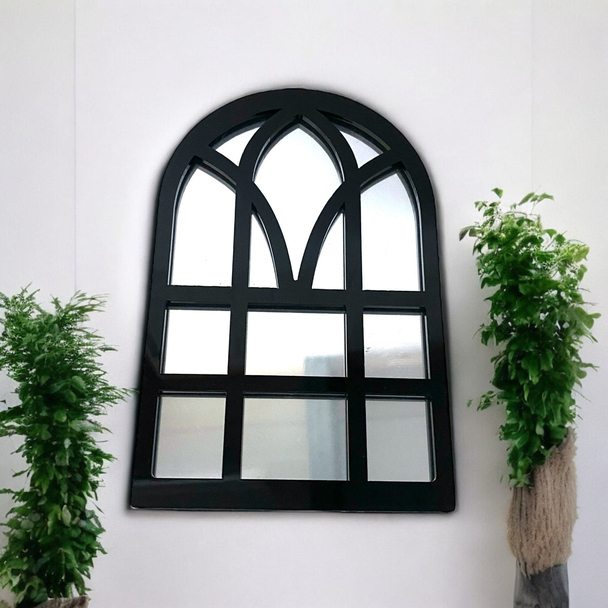 Black Victorian arch garden mirror with grid panes, flanked by two potted green plants.