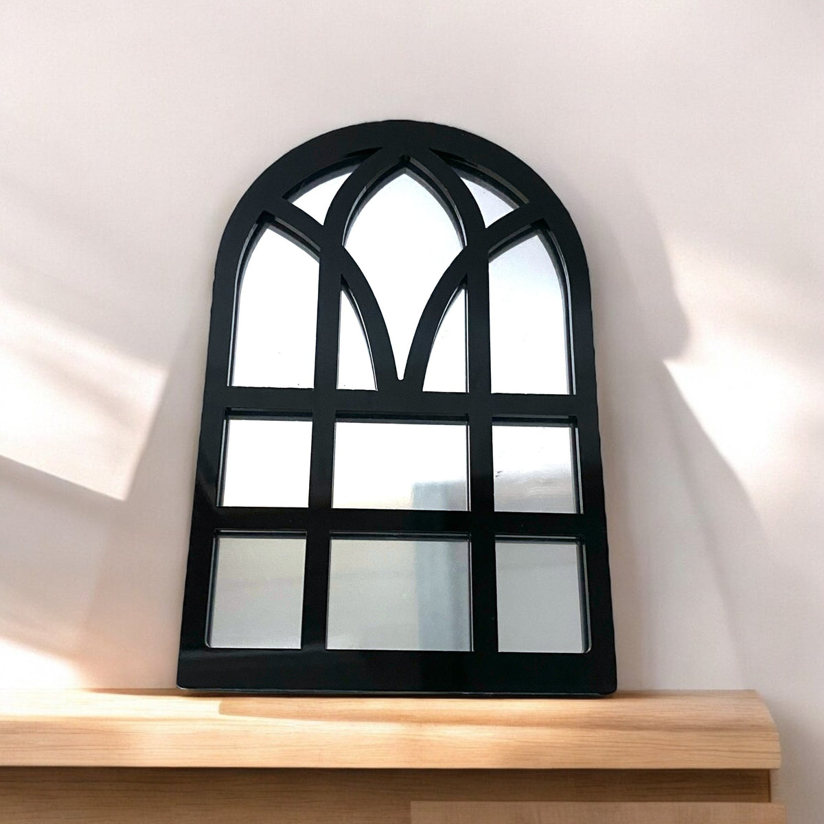 Black arched window-style mirror resting on a wooden shelf against a white wall.