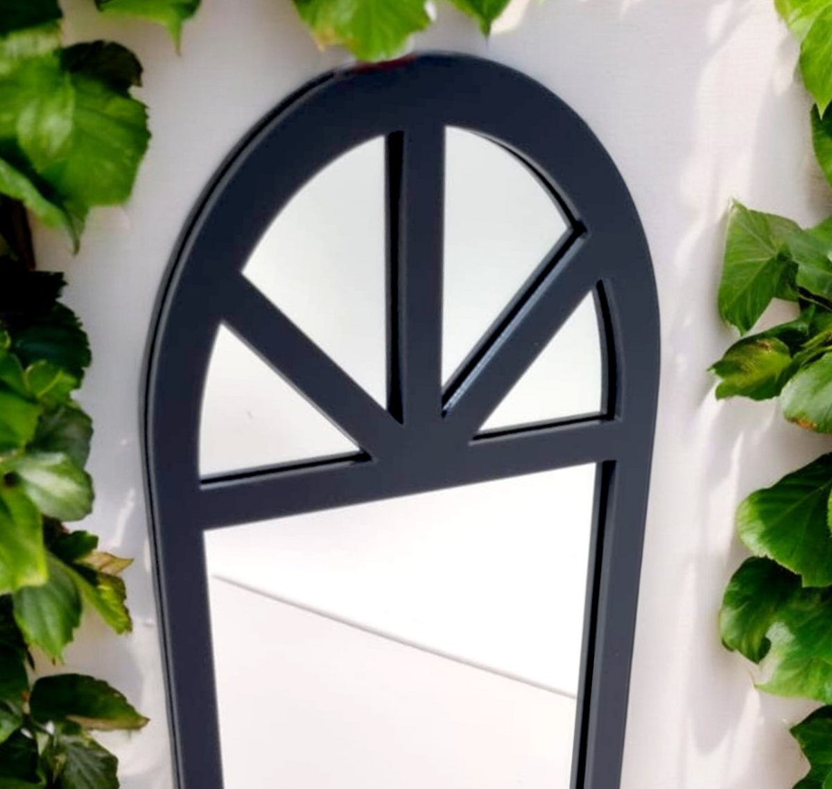 Dark gray Victorian arch framed garden mirror mounted on a white wall, flanked by green ivy.
