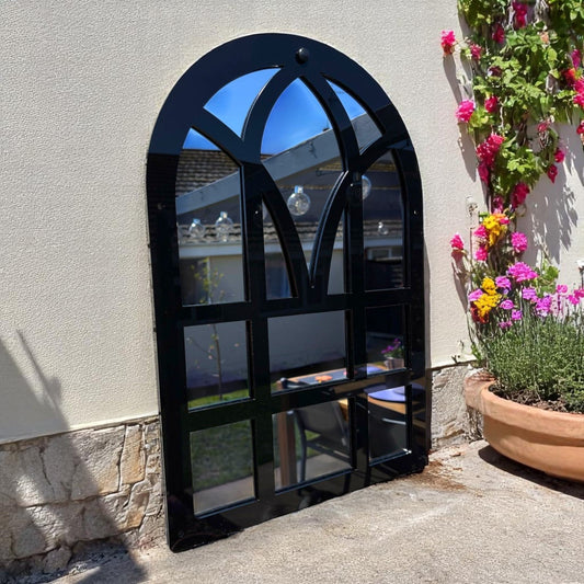 Black Victorian arch garden mirror mounted on an outdoor stucco wall with flowering plants.