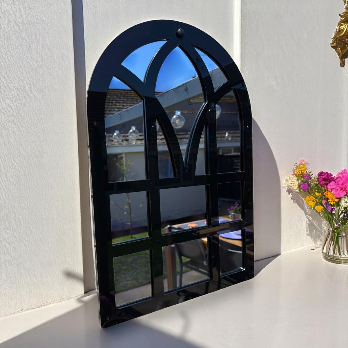 Black Victorian arch garden mirror with a windowpane frame reflecting an outdoor garden scene.