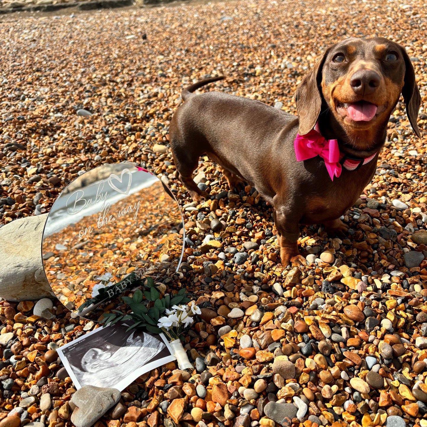 A round personalised mirror engraved with "Baby on the way" stands on a pebble beach next to an ultrasound photo.