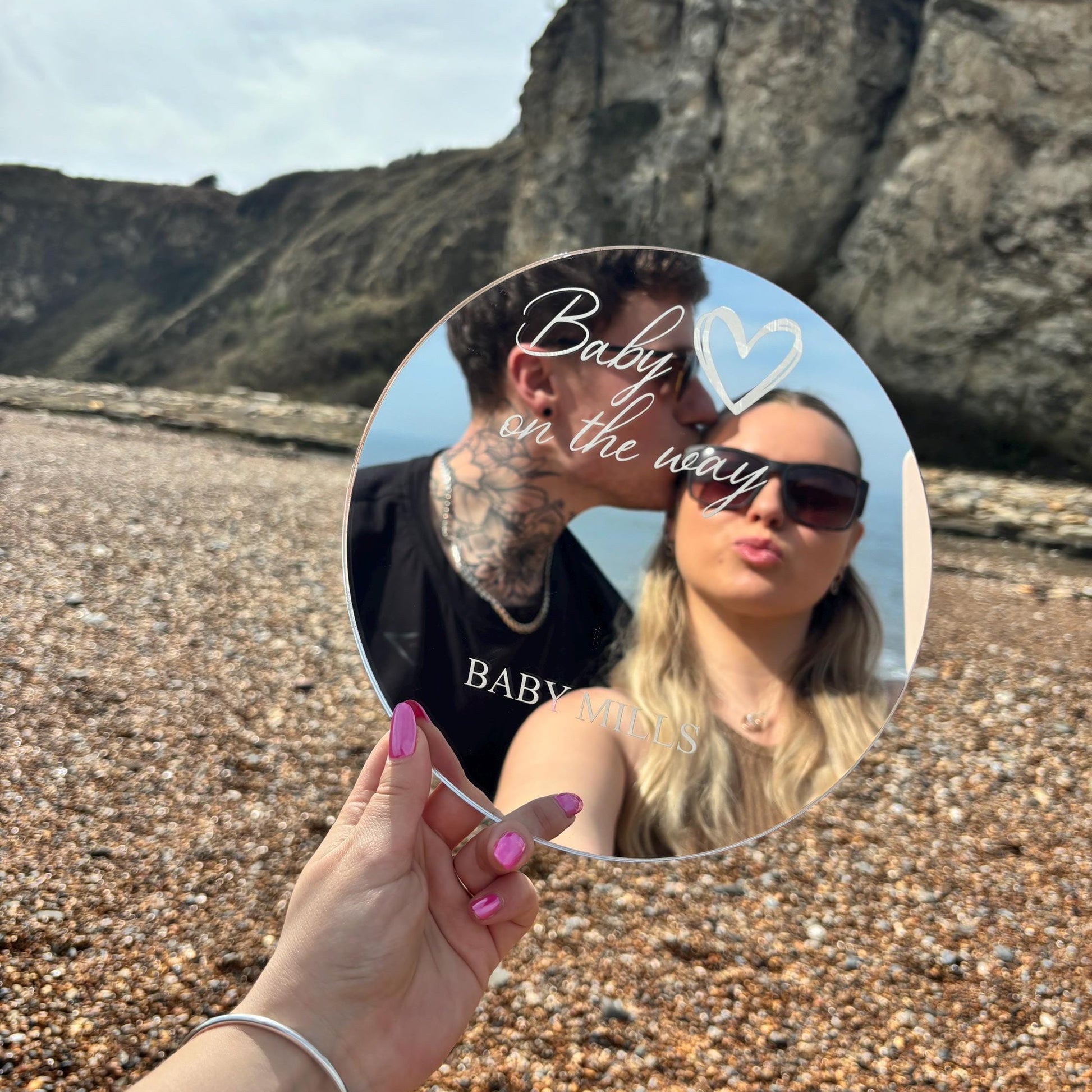 Round personalized acrylic mirror engraved with "Baby on the way" and a heart, held outdoors.