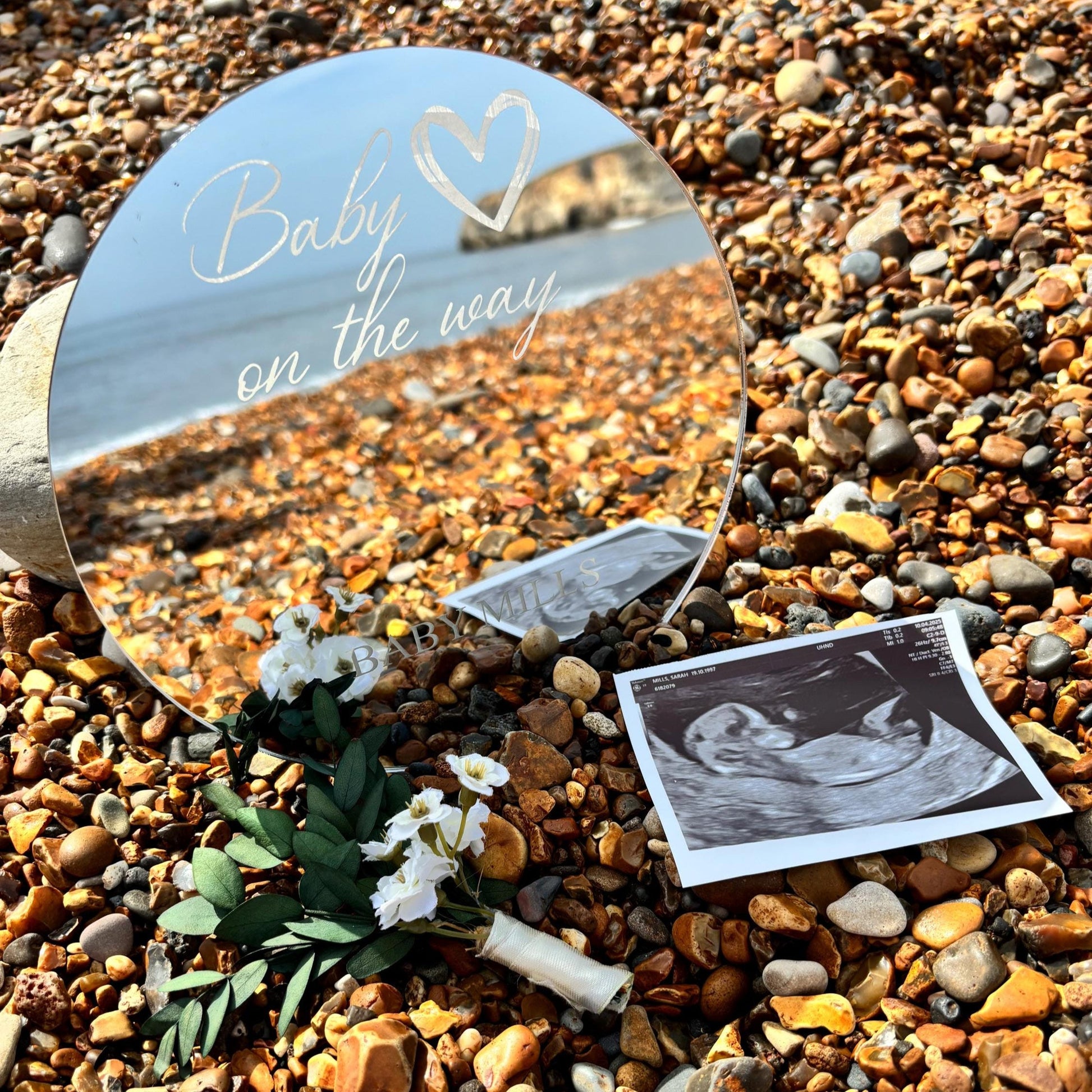 Personalised round mirror engraved with "Baby on the way" and a heart, resting on a pebble beach with an ultrasound scan.