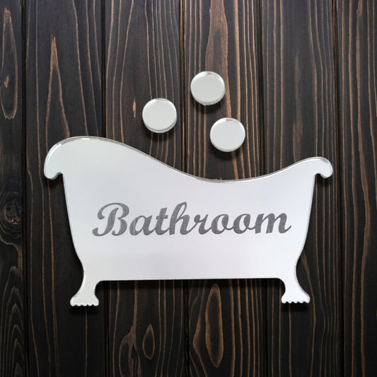 Mirrored bathtub-shaped sign with engraved text "Bathroom" and three round bubbles, on dark wood.