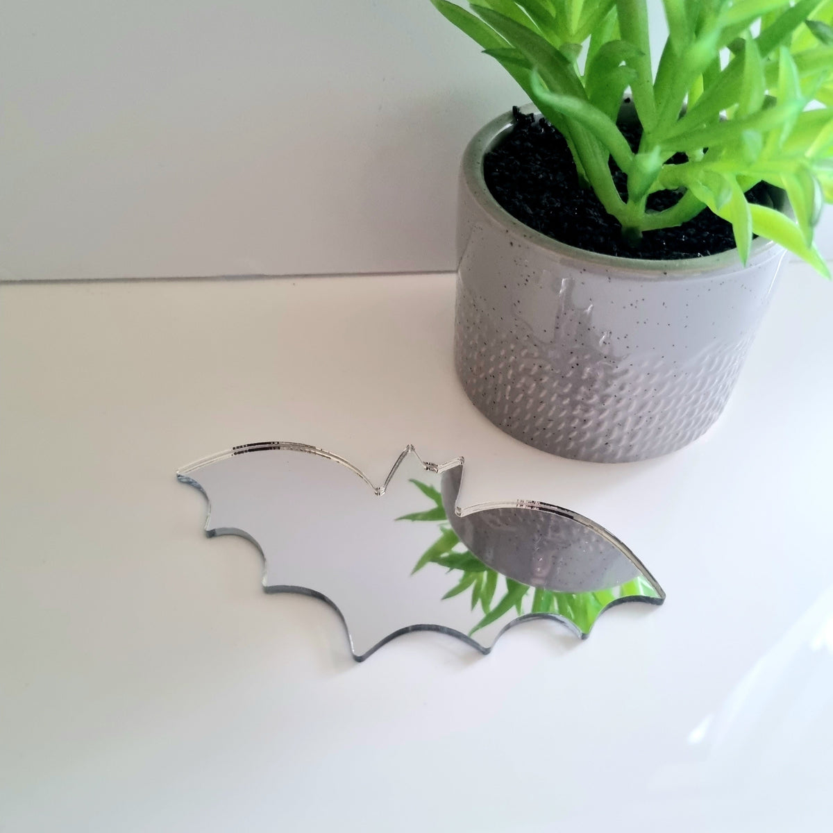 Bat-shaped silver acrylic mirror on white surface beside potted plant.