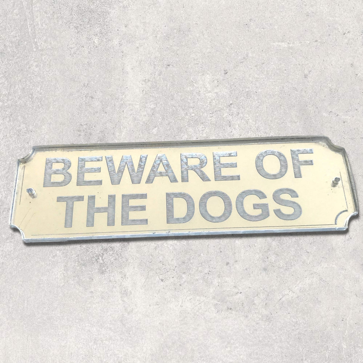 Rectangular mirror sign with silver text reading "BEWARE OF THE DOGS" on a neutral background.