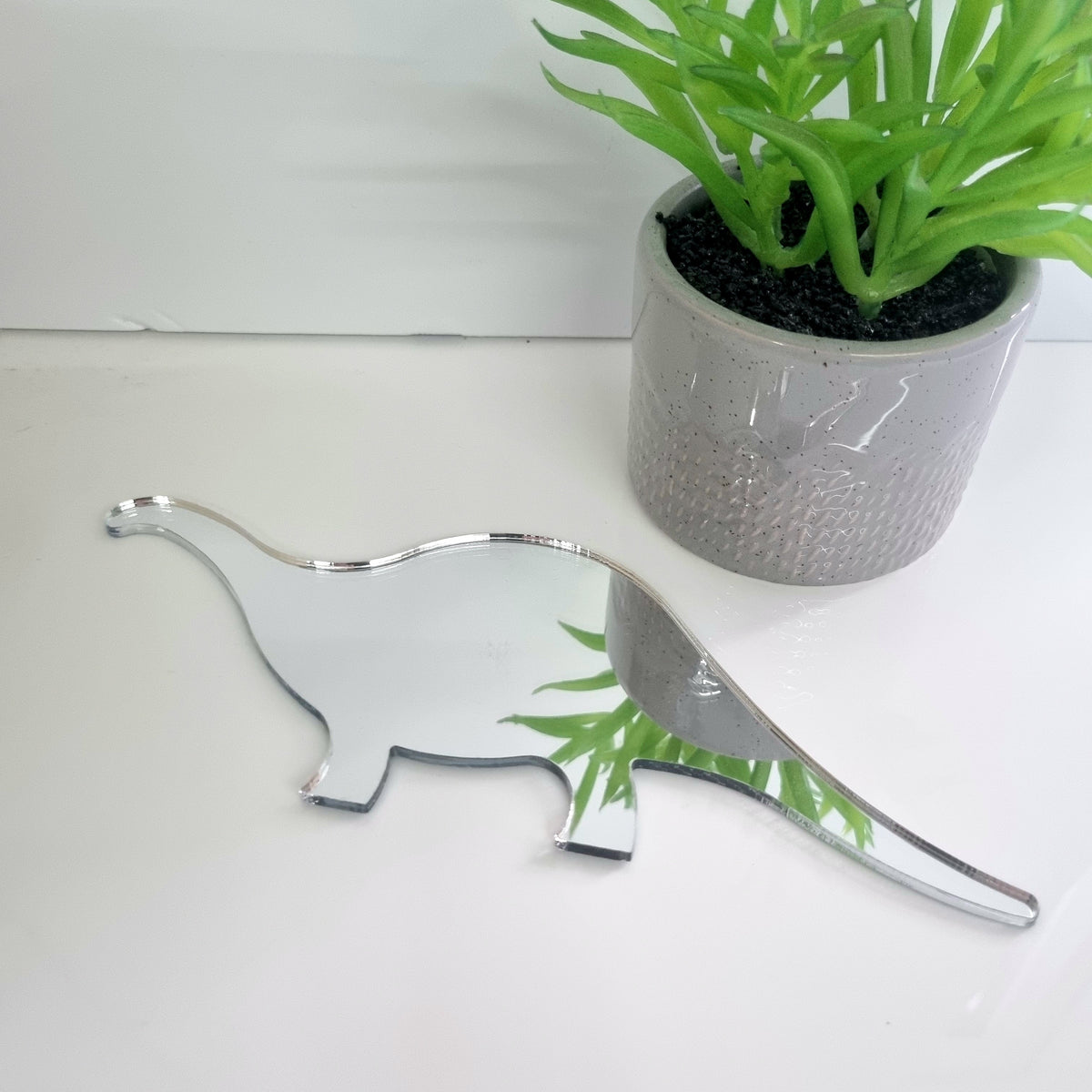 Brontosaurus-shaped silver mirror resting flat on a white surface, with a potted green plant beside it.