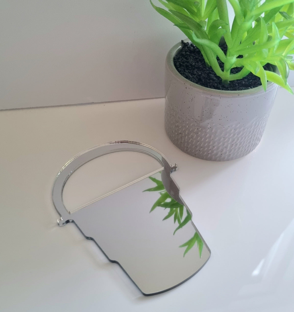 Silver bucket-shaped mirror on a white surface next to a potted green plant.
