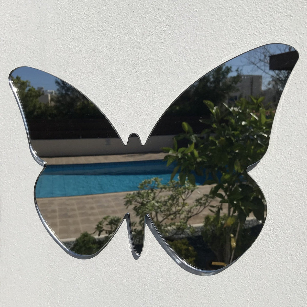 Butterfly-shaped mirror mounted on a white textured wall, reflecting an outdoor scene with a pool and plants.
