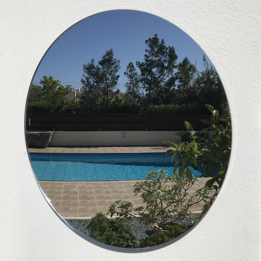 Round garden mirror mounted on a white wall, reflecting a swimming pool, trees, and blue sky.