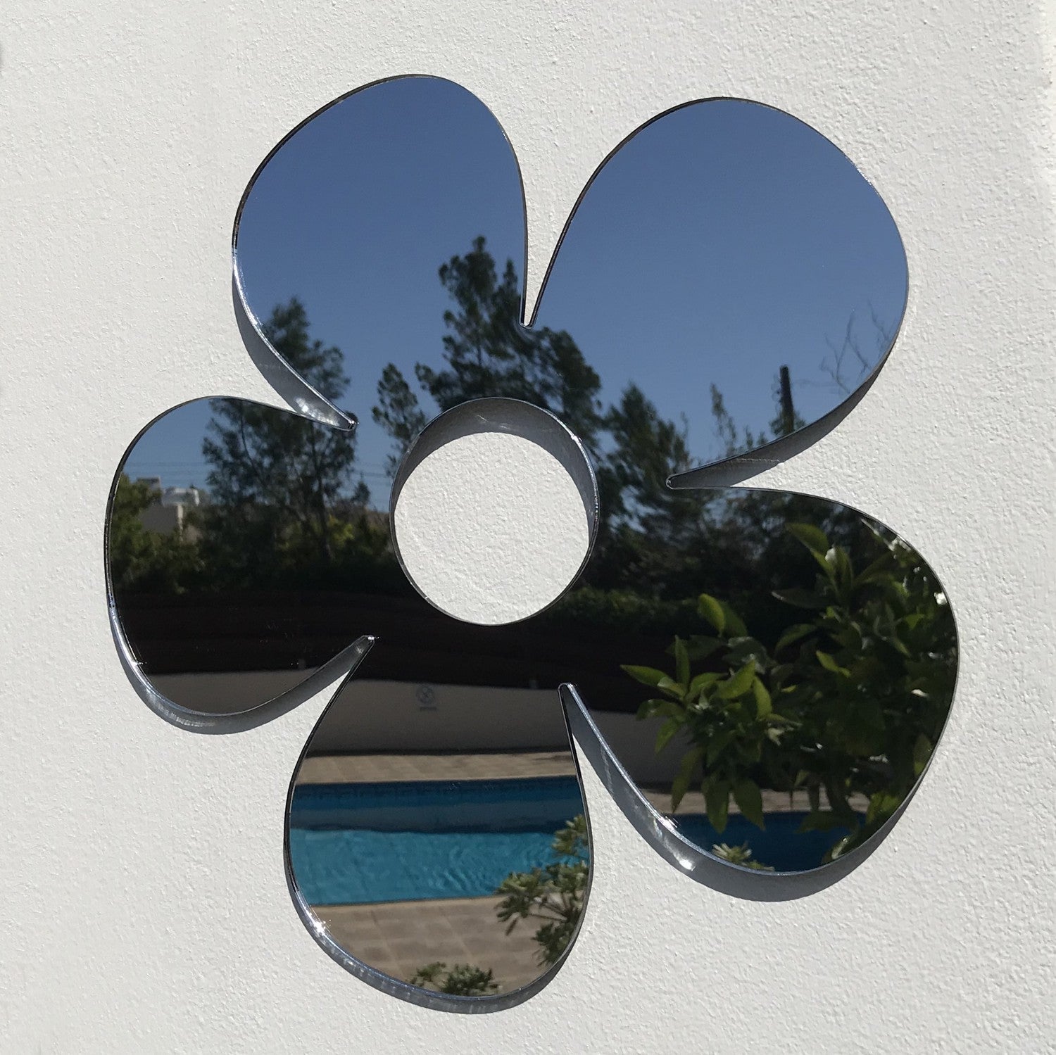 Daisy-shaped garden mirror mounted on a white wall, reflecting an outdoor pool and trees.