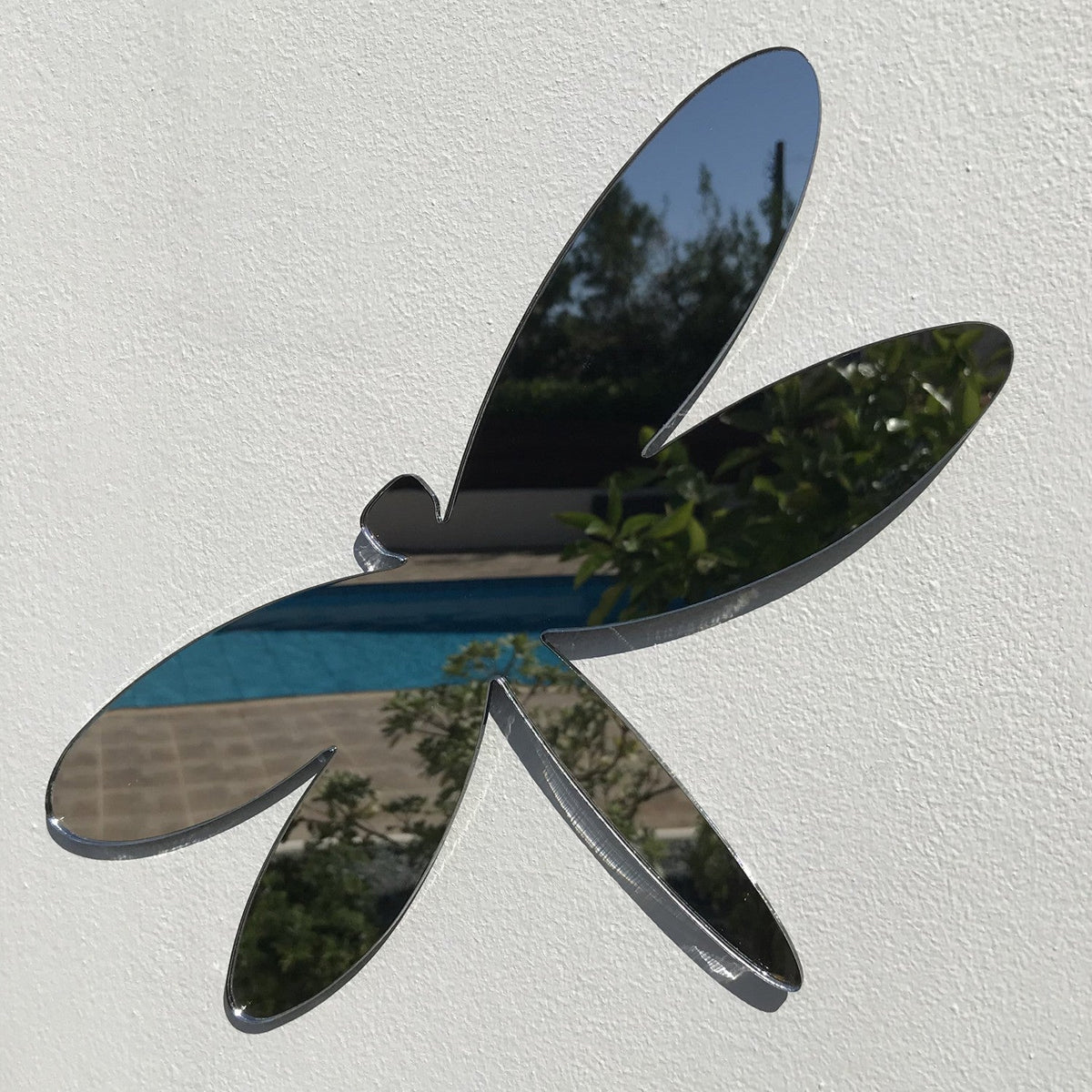 Dragonfly-shaped garden mirror mounted on a white wall, reflecting a garden scene.