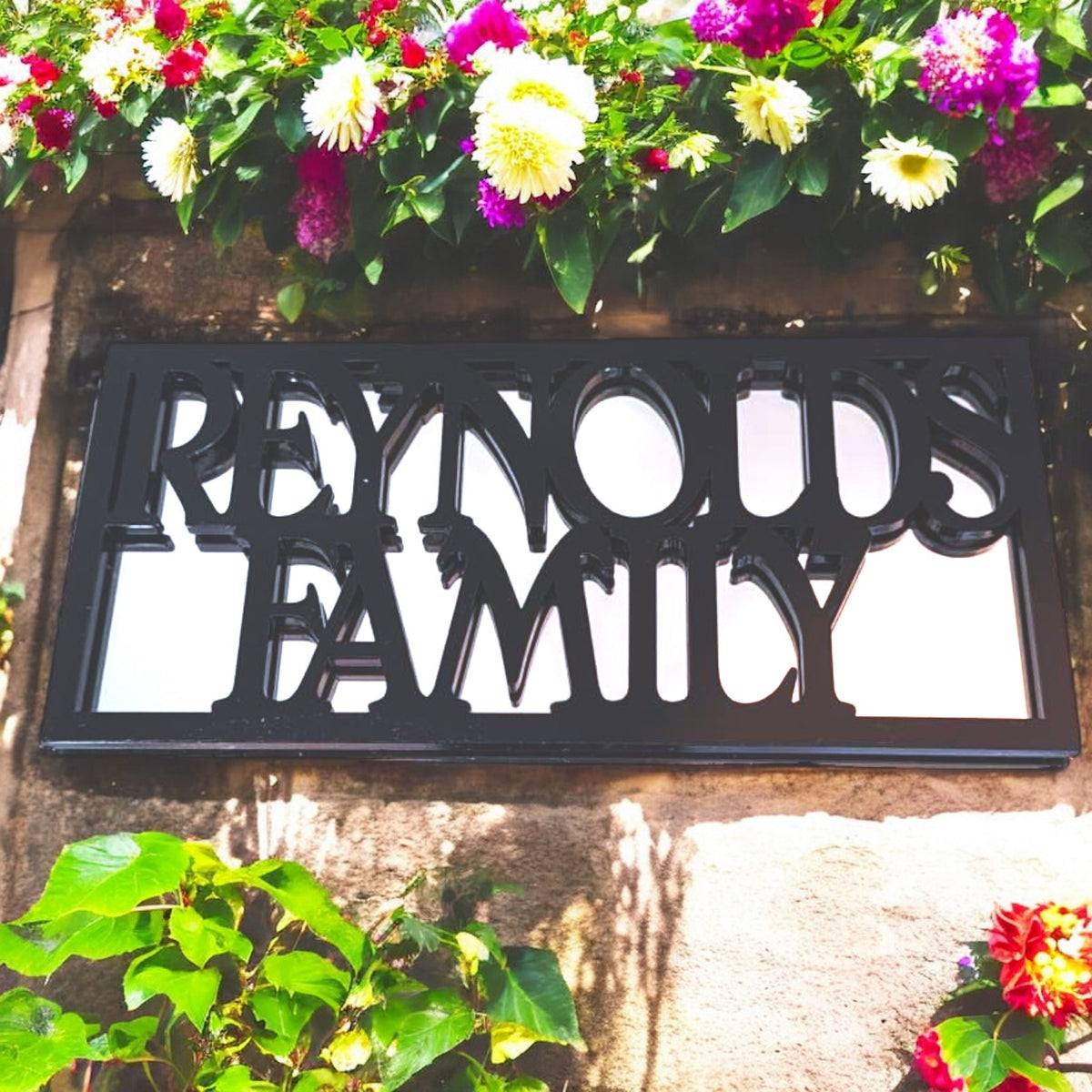 A rectangular framed garden mirror with black cut-out text reading "REYNOLDS FAMILY" mounted on a stone wall.