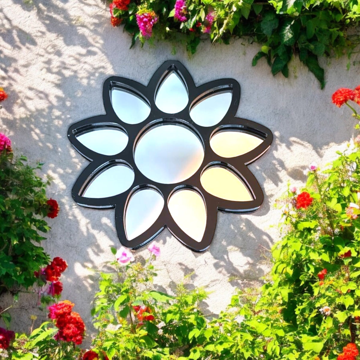 A black metal flower-shaped mirror with a round center and petal mirrors, hanging on a stone wall amid flowers.