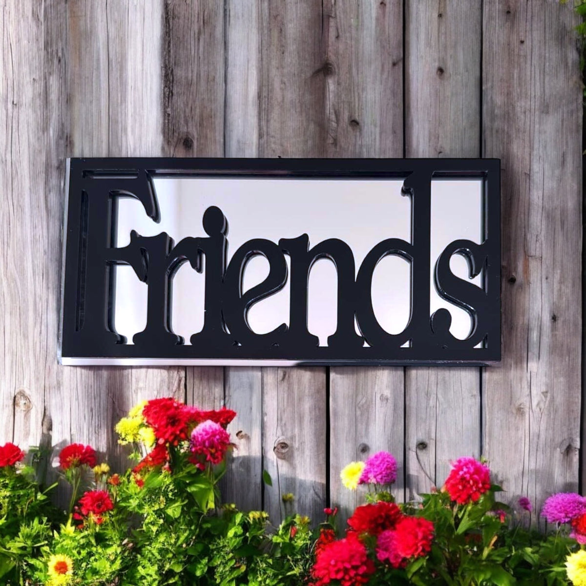 Black rectangular framed garden mirror featuring the cutout word "Friends" mounted on a wooden fence.
