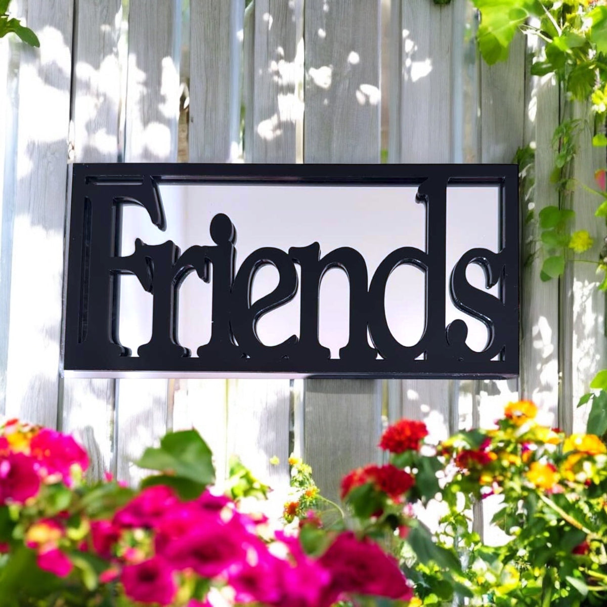 Rectangular black metal framed garden mirror reading Friends mounted on a wooden fence.