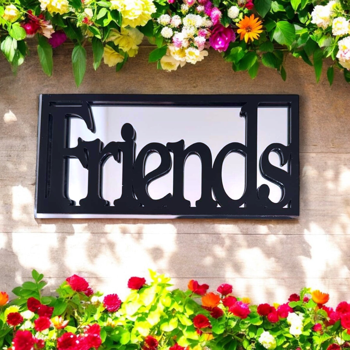 Black-framed rectangular mirror featuring cutout "Friends" lettering, mounted on a wooden wall with flowers.