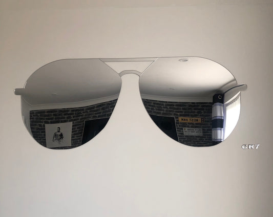 A large silver-framed mirror shaped like aviator sunglasses mounted on a white wall.