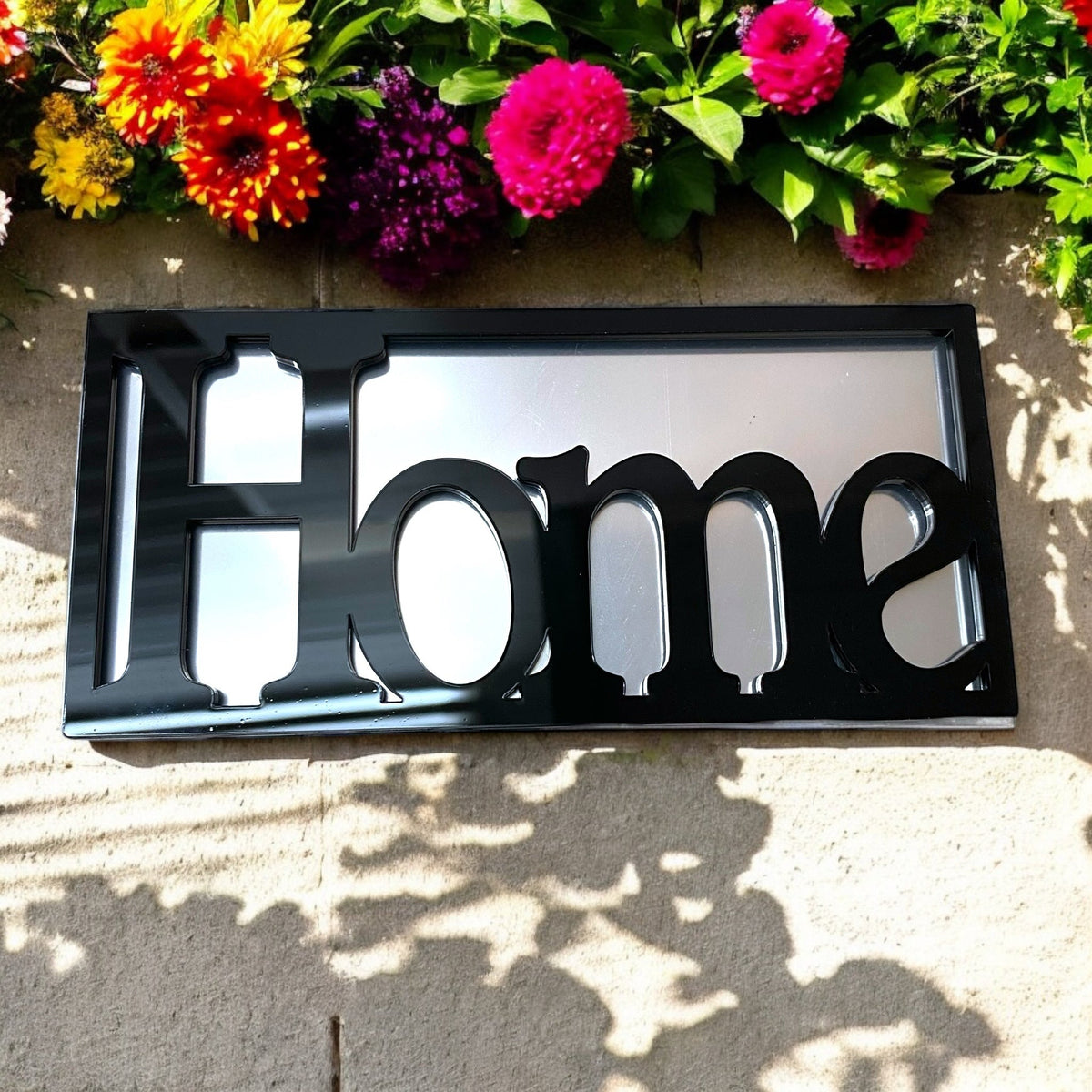 Black rectangular framed mirror featuring the cut-out word "Home" on a stone wall.