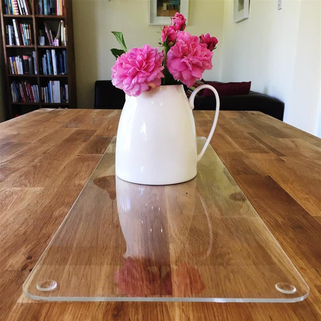 Clear, rectangular acrylic table runner protector on a wood table, holding a white pitcher with pink roses.
