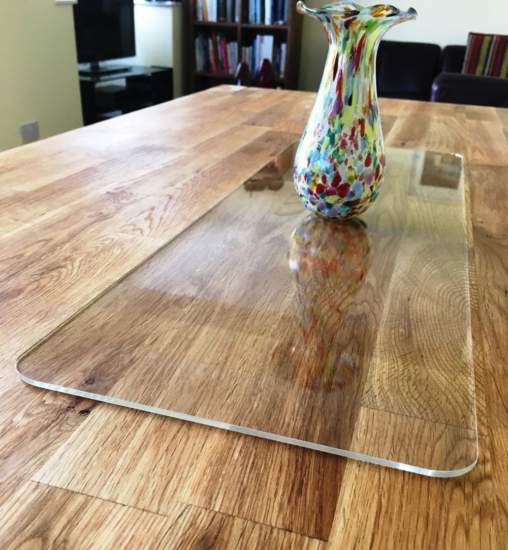 Clear rectangular acrylic table runner with rounded corners resting on a wooden table, topped with a colorful vase.