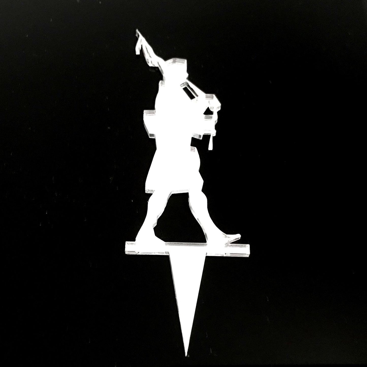 Clear acrylic cake topper silhouette of a Scottish bagpipe player on a pointed stake against a black background.