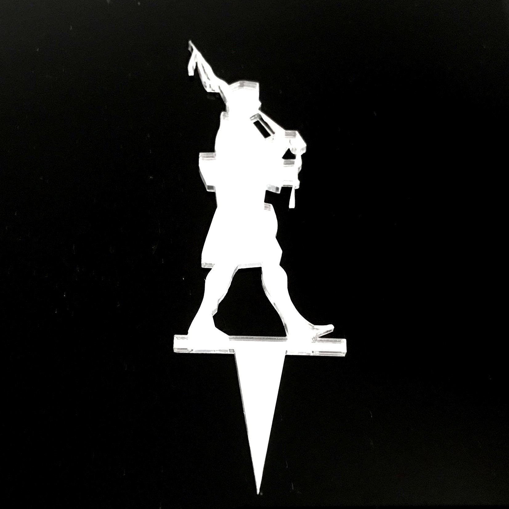 Clear acrylic cake topper silhouette of a Scottish bagpipe player on a pointed stake against a black background.