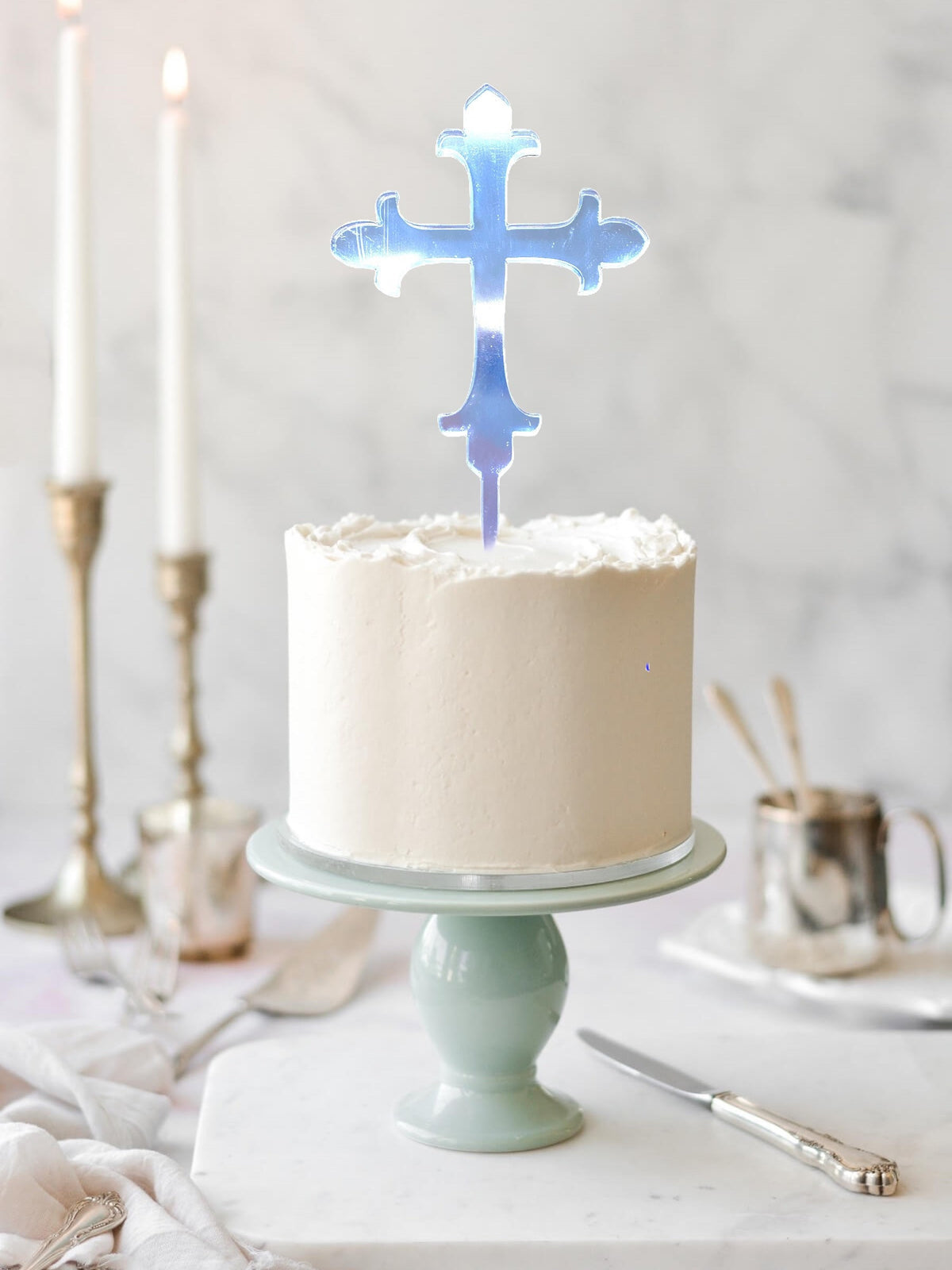 Iridescent, laser-cut acrylic cross cake topper on a white frosted cake on a pastel green pedestal stand.