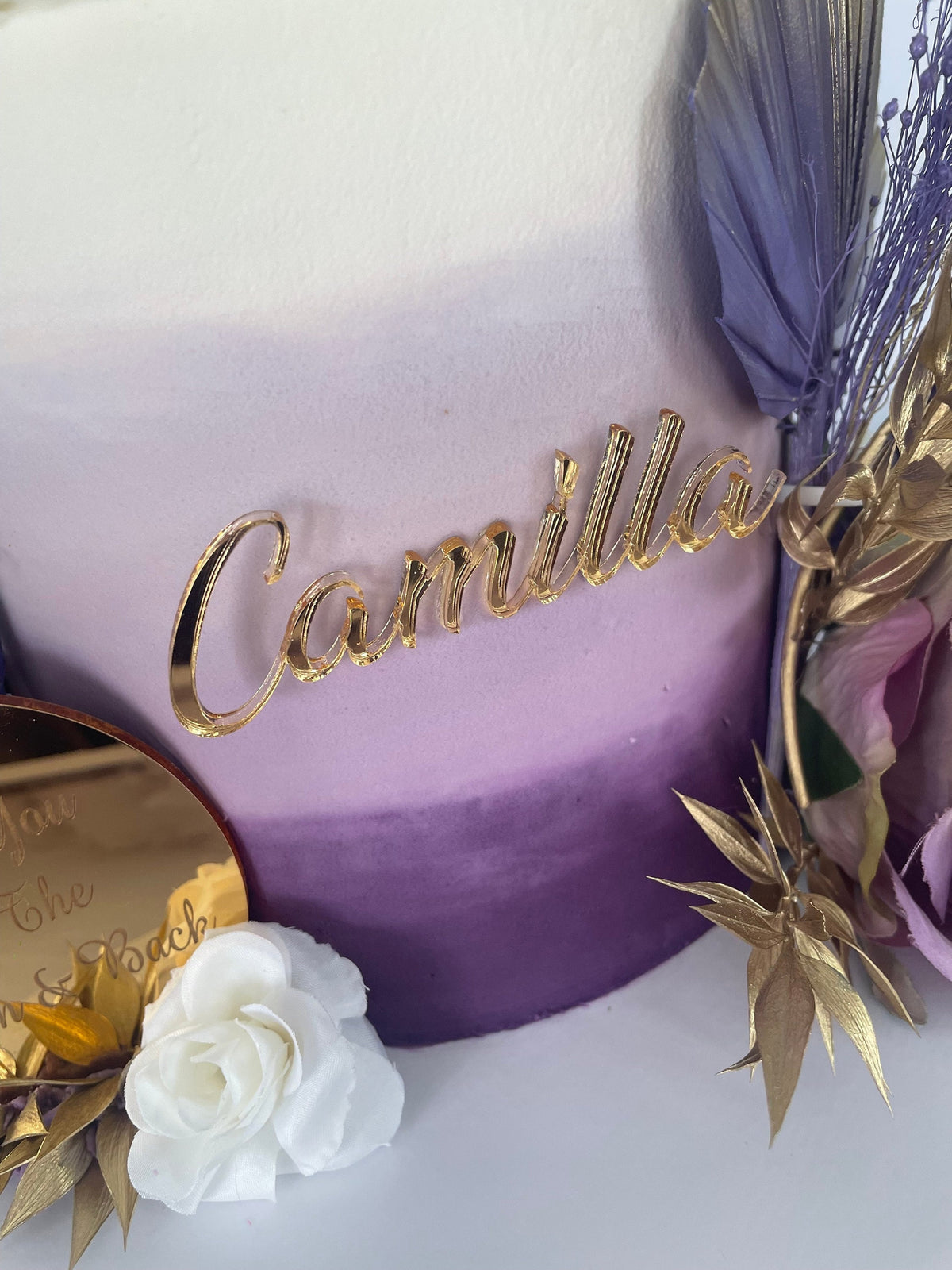 Gold acrylic, cursive "Camilla" cake topper attached to a purple ombre cake with floral decorations.