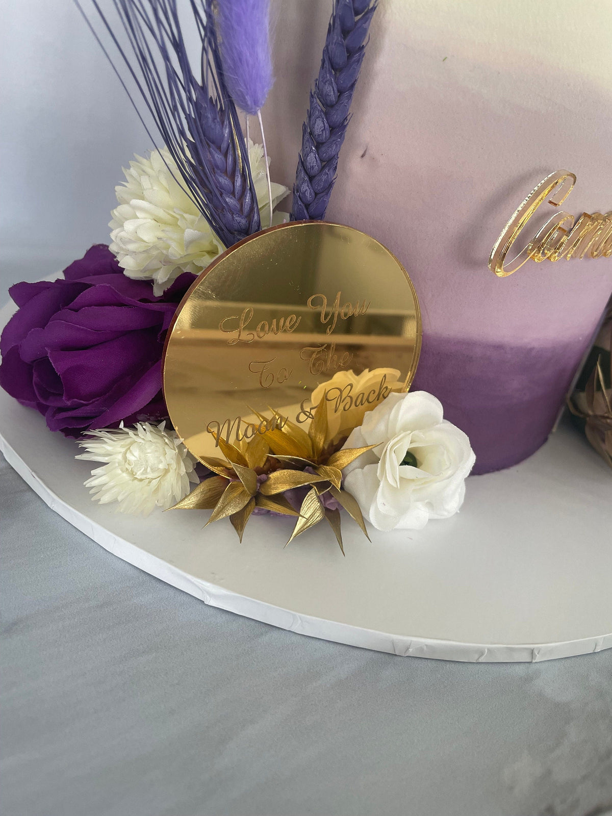 Circular gold mirror acrylic cake topper engraved with "Love You To The Moon & Back," nestled in purple floral decor.