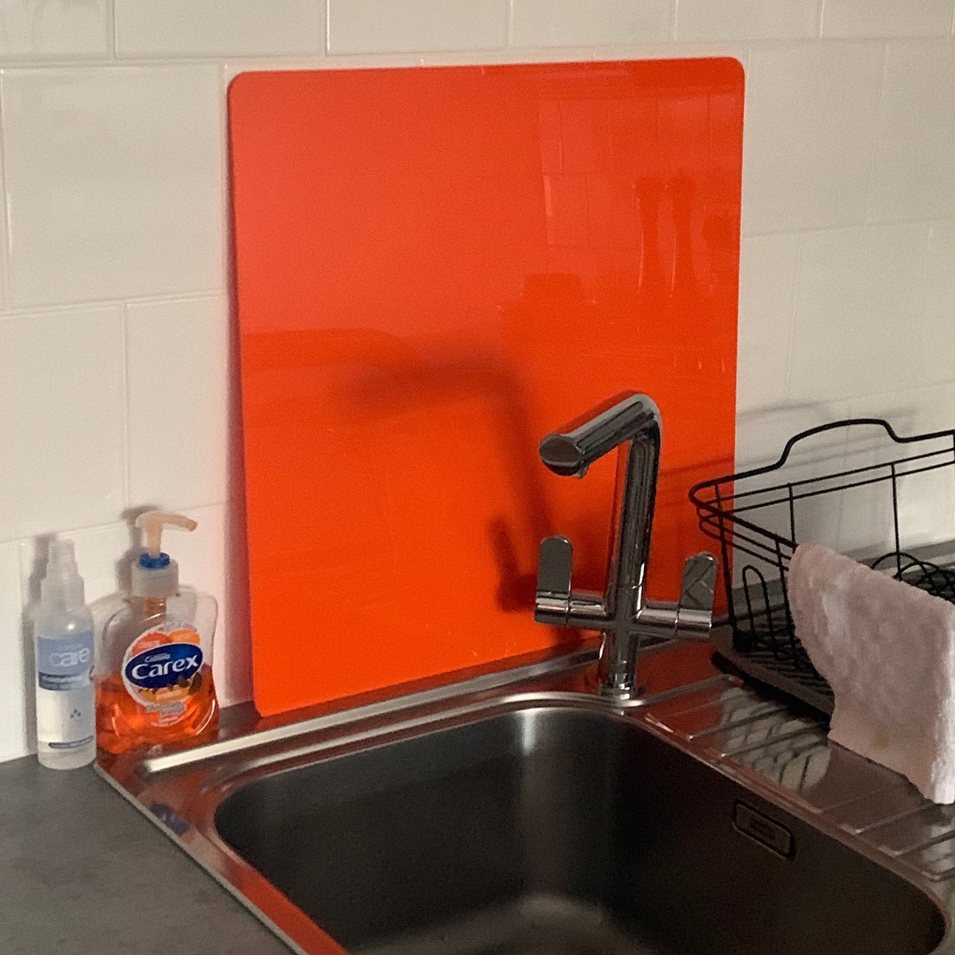 A vibrant, square orange acrylic worktop saver standing behind a kitchen sink and chrome faucet.
