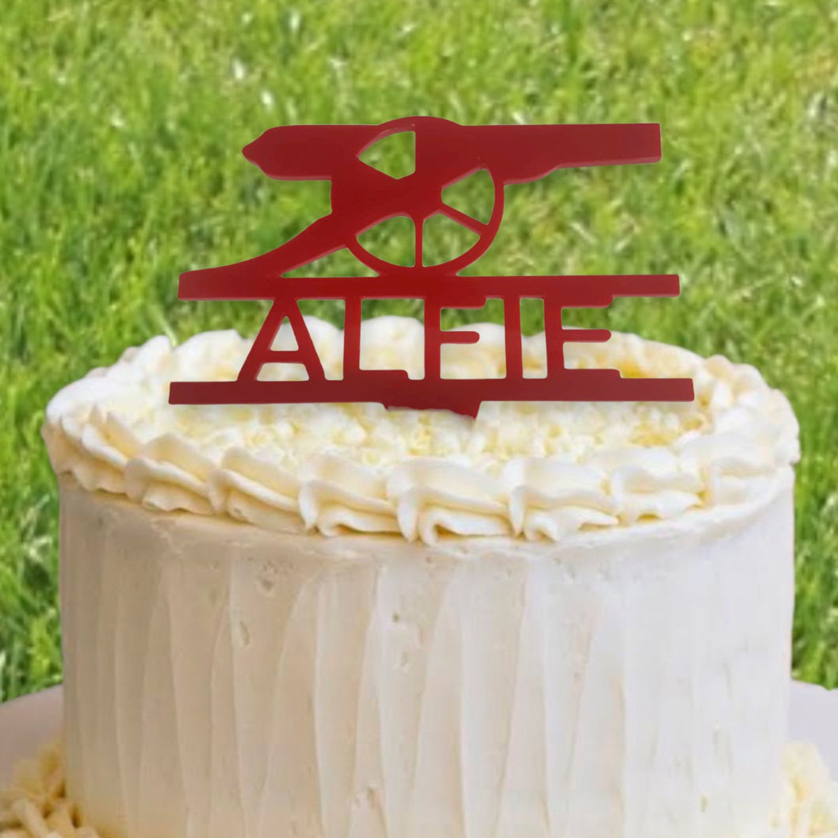 Red acrylic custom cake topper featuring the Arsenal FC cannon logo with the name "ALFIE" on a frosted cake.