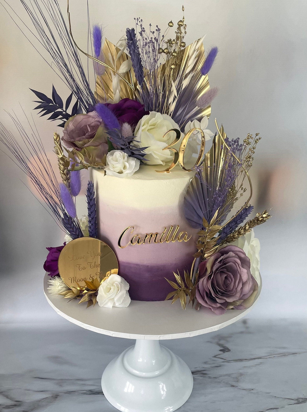 Gold "Camilla" and "30" acrylic cake toppers on an ombre purple cake decorated with dried flowers and gold palms.