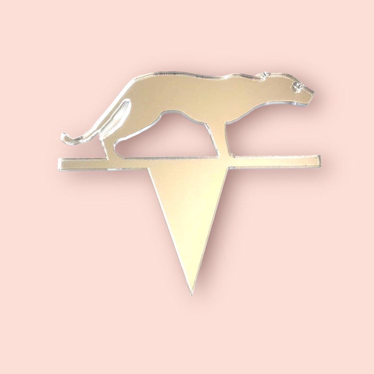 A mirrored gold acrylic jaguar silhouette cake topper with a triangular spike base on a pale pink background.
