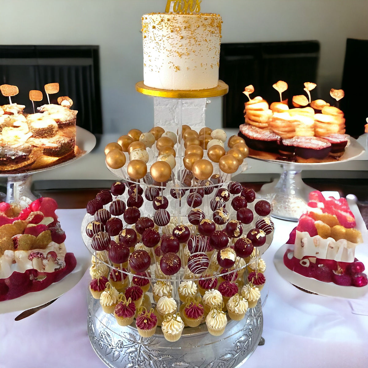 Clear acrylic tiered cake pop stand by SuperCoolCreations holding gold, maroon, and white cake pops and mini cupcakes.