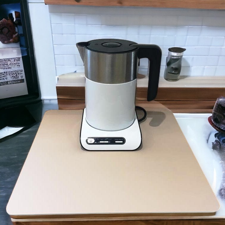 A square, matte beige acrylic worktop saver under a stainless steel and white electric kettle on a wooden counter.